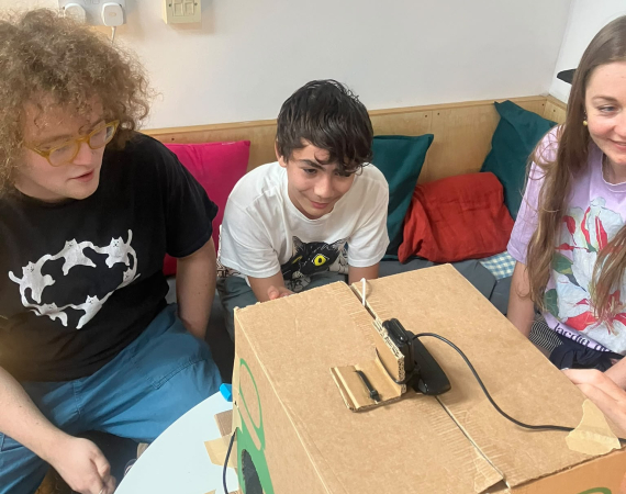 Three young people gather around a large cardboard box with wires and a handle, appearing curious and engaged in a colorful indoor room.