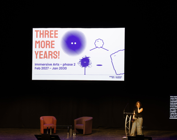 Speaker stands on a dimly lit stage beside a slide reading “THREE MORE YEARS!” with colorful drawings, creating an optimistic, creative tone.
