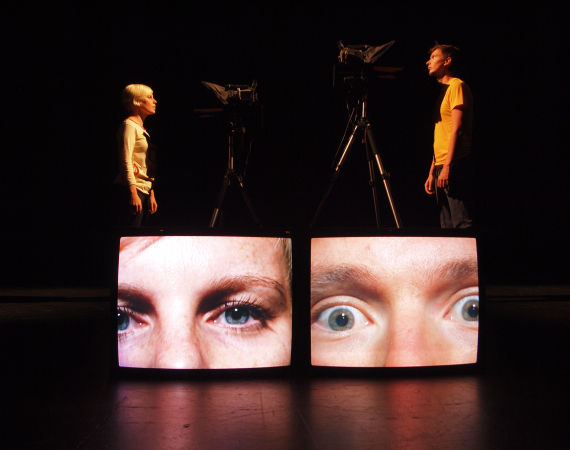 Two artists stare at each other, infront of them are close up projections of their faces on two monitors.