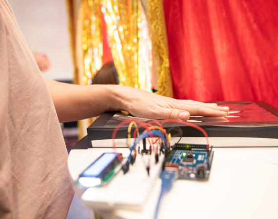 Image of wires, a circuit board and a persons hand on a tablet. There is red and gold fabric in the background.