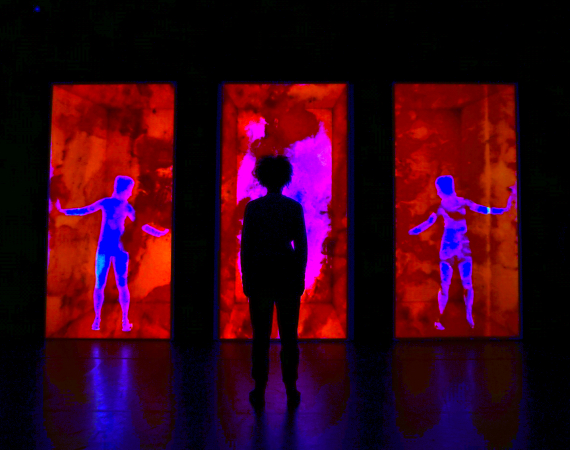 A person stands silhoutted infront of 3 red screens. The screens feature body shapes on them.