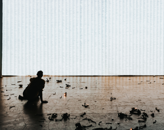 Man sitting on the floor with his back turned, looking at the white empty wall with some bones, burnt pieces of set on the floor