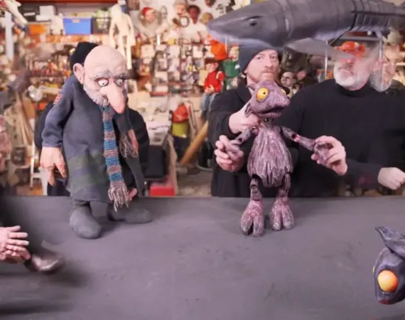 two puppets stand on a table. 3 of the puppeteers are visible behind them.