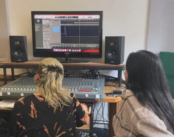 Two people sit infront of a desk with a monitor and a sound desk.