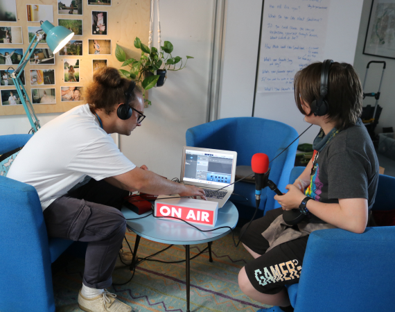 Two people sitting in front of a laptop with microphones and an 'on air' sign