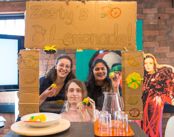 Three people are excitedly in the middle of a frame reading, 'Zesty's Lemonade' all holding lemons.