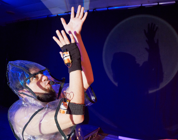 A young white man posed with his arms in the air and is wearing a see through raincoat with batteries attached to him and black fingerless gloves