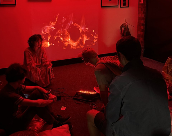 A group of people sit in a dimly lit room with red lighting, focused on a vibrant fire projection on the wall. Modern art hangs above them.