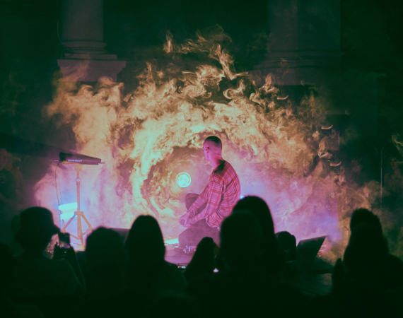 Performer holding a microphone on a foggy, dimly lit stage with an audience silhouette in the foreground.