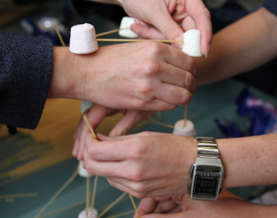 hands coming in from all directions, building something out of spaghetti and marshmallows