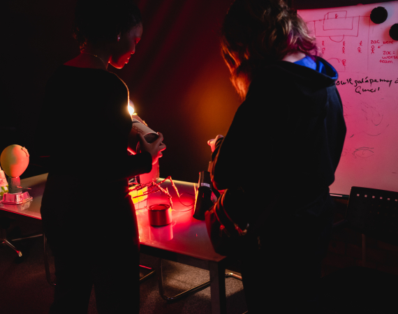 Two people examine a small object at a cluttered worktable under red light, beside a whiteboard covered in sketches and notes; the mood is focused and tense.