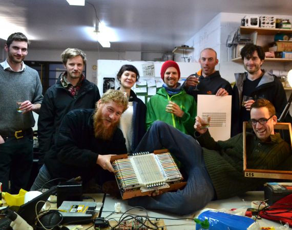The Canute team posing for the camera with a partially assembled Canute Mk8 and glasses of whisky in a busy workshop.