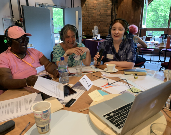 Three women with headphones speaking to someone on a laptop and laughing. There is lots of paper the table they ate sat around.