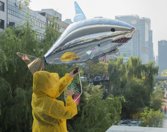 A person wearing a yellow raincoat with the hood up holding a silver shark balloon. The person is standing in front of green trees with many tall buildings in the background. To the right of the image is a path going in the city, it has a number of people walking.  