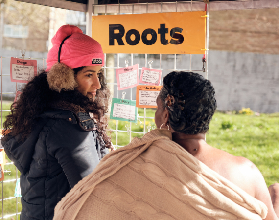 Play:Disrupt Creative High Streets Project, two people outside looking at note cards under an orange sign saying 'roots'