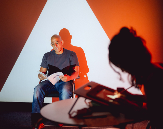 A man is reading from a book sitting in front of a wall where live illustrations are projected. The live illustrator is out of focus in the foreground.