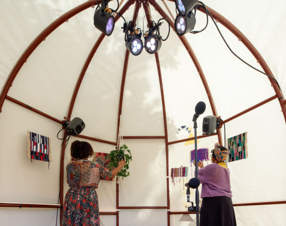 An image of the inside of a domed house with curved iron ribs onto which white canvas has been tautly wrapped around. Traditional Somali prints on the outside of the canvas are also visible from the inside of the house. We can see the backs of two women facing various colourful small woven structures, and a potted plant, all hanging from the ribs of the house. A microphone and lights are also visible inside the space.