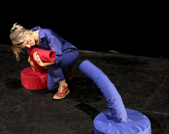 A person in a blue boilersuit is hugging a soft red tubular sculpture facing a similar looking blue sculpture. This is in a black box theatre stage. 