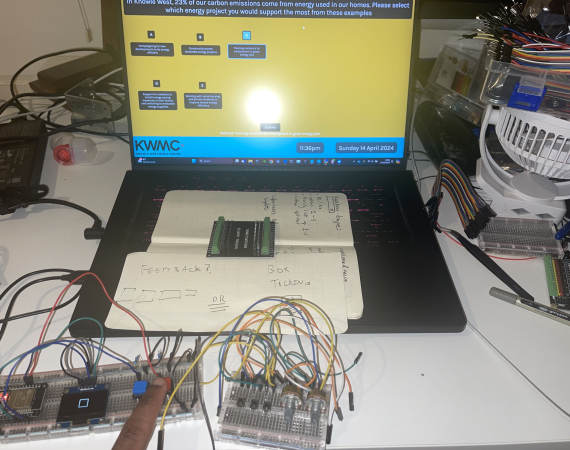 Open laptop with some control boards in front of it and notes. A person's finger is pressing one of the electronic buttons.
