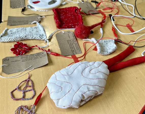 A selection of red and white electronic textile sample on a beige/brown table. The samples include knitting, fabric and felted fibres and each is plugged in my a crocodile clip and has a brown tag to identify it.