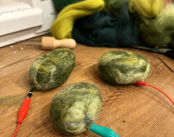 Three green pebble-shaped felted balls with a crocodile clip attached to each. In the background there is a pile of green felting fibre and the corner of a sewing machine