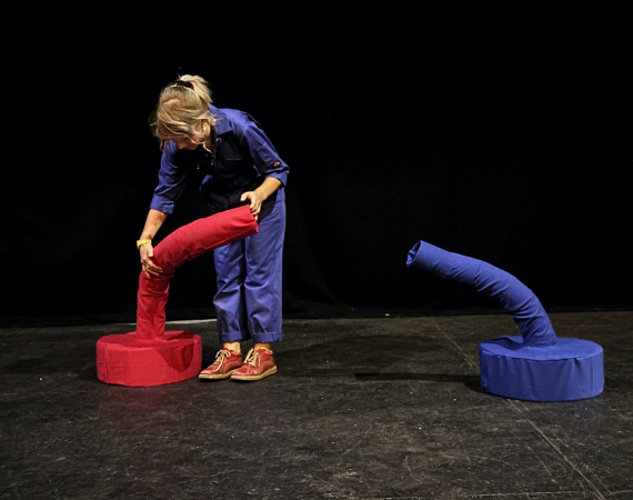 A person in a blue boilersuit is hugging a soft red tubular sculpture facing a similar looking blue sculpture. This is in a black box theatre stage.