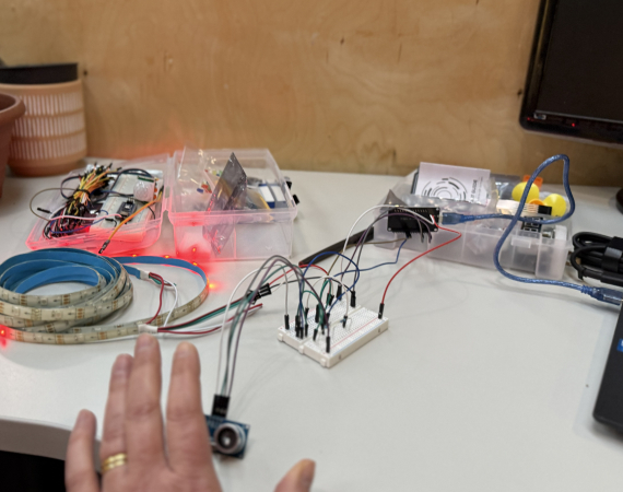 A hand waves in front of a sensor connected by electrical wires via a breadboard to an LED strip and an Arduino