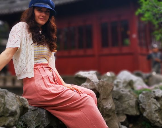 profile picture of Ellie Chadwick: woman in early 30s, sitting on a rock in front of a pagoda, with long dark hair, a blue hat, yellow striped top and pink trousers