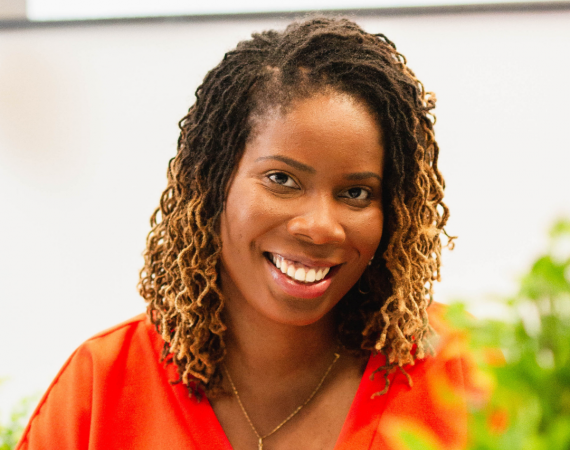 Joyann is smiling looking directly to the camera, she has should length locs and is wearing a orange dress