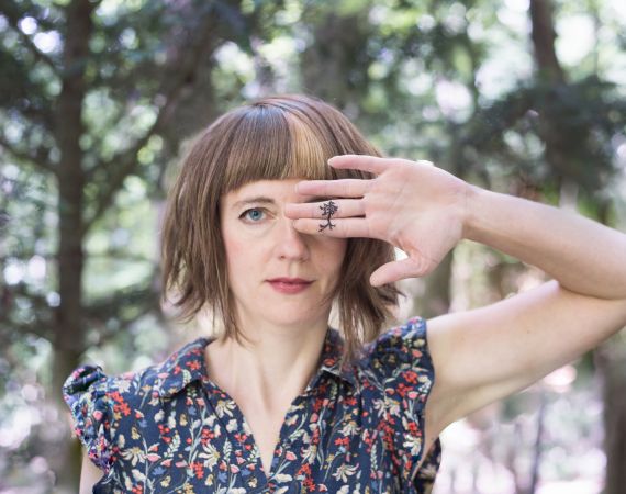 A close-up of artist Sylvia Rimat in a forest, with the palm of one of her hands over one of her eyes, revealing a tattoe-like drawing of what could be a tree or a neuron.