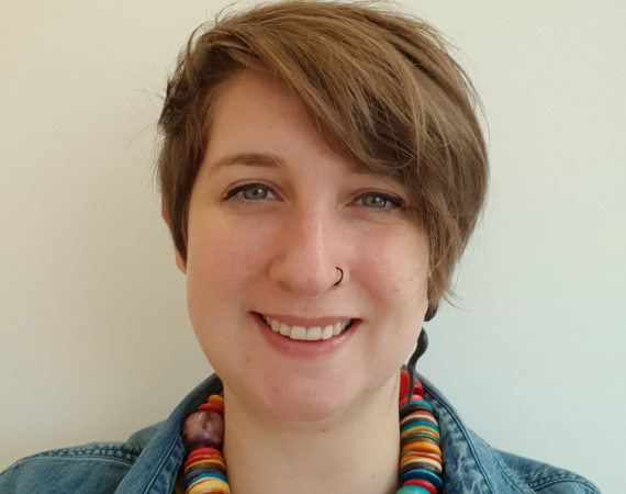 white women with short brown hair. Wearing a denim shirt and multi-coloured necklace.
