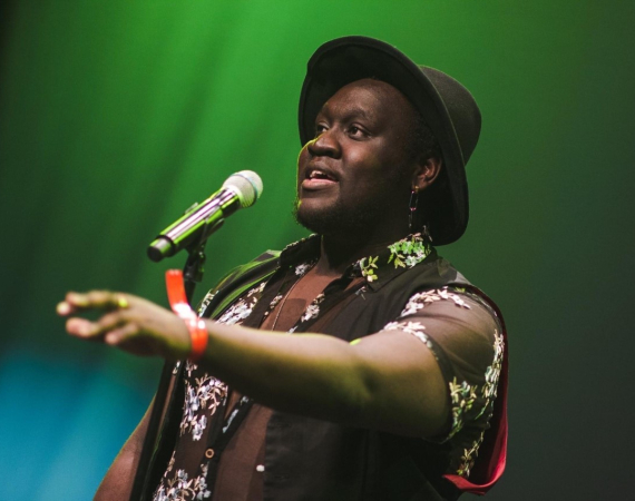 Imagine of a Black man on stage performing. The background has a green light and he is wearing a black shirt with a wide-brim hat speaking into a microphone