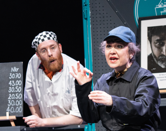A colour photograph of a white man in his 30s with a short ginger beard and moustache wearing a black and white checkered chef's cap, and chef's whites. He's leaning over a large grey panel that serves as a serving hatch in a cafe. In front of the serving hatch is a white woman in her 40s wearing a black cap and a black boilersuit. She's miming holding a cup and the other hand signals the OK sign for divers, fingers forming a loop with the little fingers sticking up. 