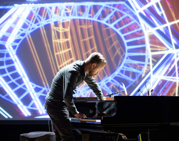 Larkhall reaches inside a piano while colourful visuals are projected behind him