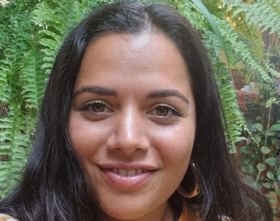 A brown woman with long dark hair and big smile. Roseanna wears a bright orange jumper and is standing in front of a lush green plant.