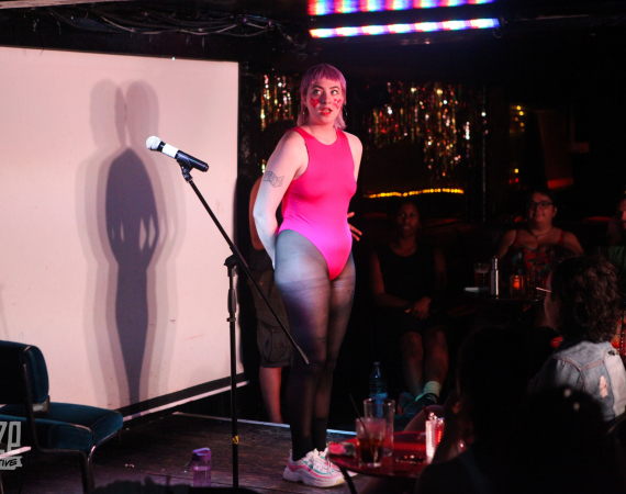 Raina is a white nonbinary person with short pink hair. They are standing on stage next to a mic and projection screen. She is wearing an 80's style pink leotard, black tights, and had pink hearts drawn on her cheeks. They are looking slightly surprised, to the left of the audience.