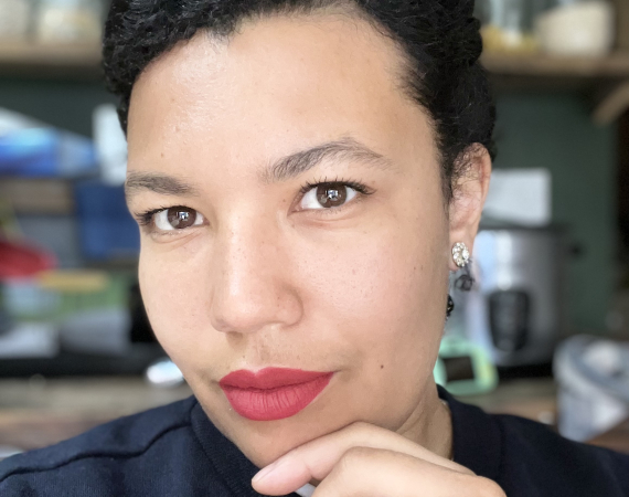 Portrait picture of a Jamaican/Scottish woman slightly smiling with bright red lipstick. Her black curly hair tied loosely back, her chin resting on her hand
