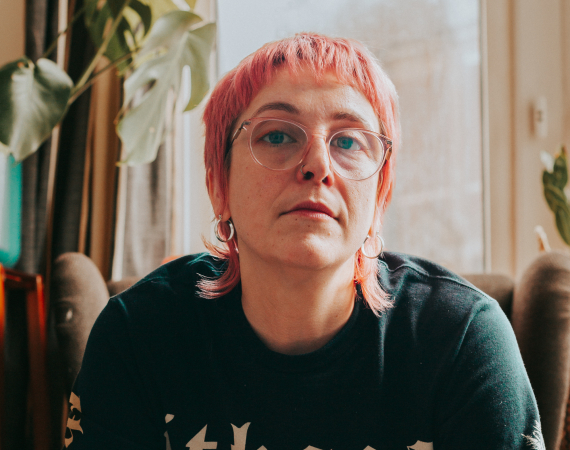 Portrait shot of Bridget in colour, with pink hair and glasses, wearing a black shirt in front of a window