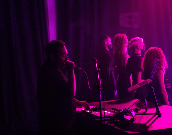 Artist Nik Rawlings, dressed in black, sings into a microphone behind a group of performers who are looking away from them, while they're all washed in pink light.
