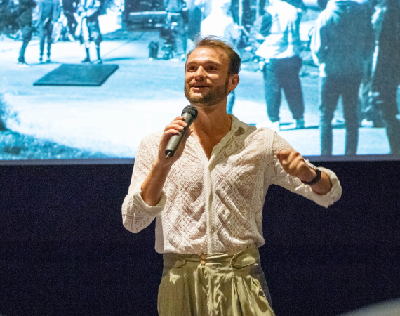 White male in a white lace shirt presenting in front of a cinema screen.