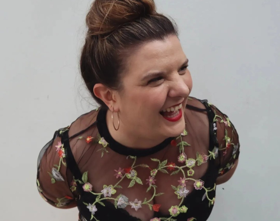 Liz, a white woman with brown hair tied in a bun is looking to her left and laughing at something off-camera, she wearing a black and embroidered flowery top and her staple red lipstick!