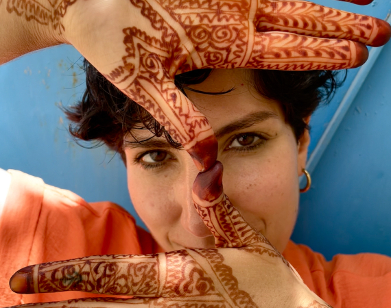 Leila against a bright blue door, her face in the centre of the image, wearing a bright orange shirt, gold hoop earrings, and black kohl eyeliner. She has short dark hair, and is looking directly at the camera, holding up both of her hands in front of her face - one with her palm-side facing the camera and one with her front-side. Her thumbs are touching at the tips of her fingertips in the centre of the image, and her hands are covered in intricate deep orange henna designs that extend onto her wrists