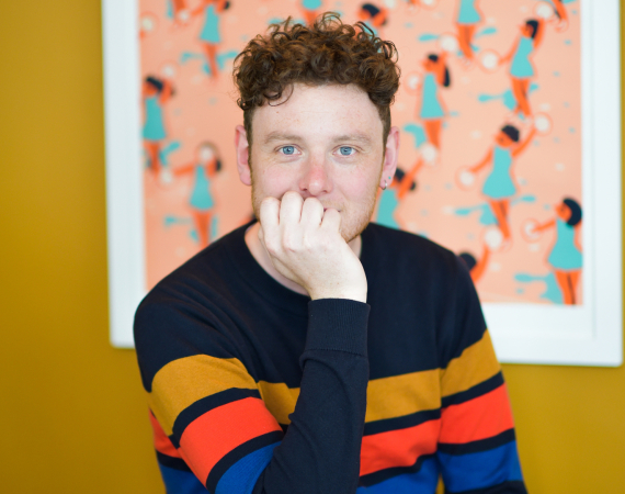 Jon is a white man with brown, curly hair. He is smiling, with his hand partially covering his mouth. He is wearing a dark jumper with three colourful stripes. He is sat behind a table looking into the camera, with colourful artwork on the wall behind him.