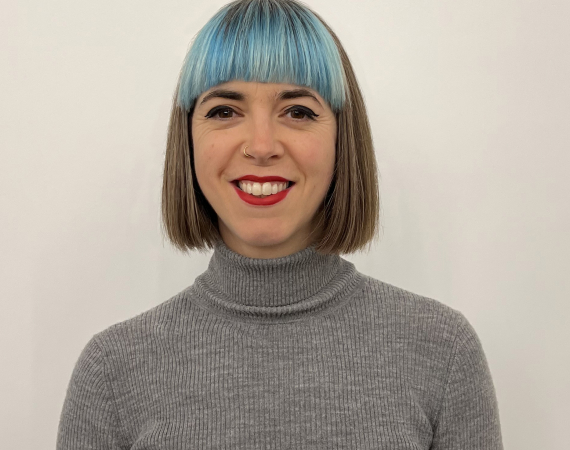 Portrait of Gabby Shiner-Hill, a smiling woman with a straight bob, blue fringe and a grey turtleneck top.