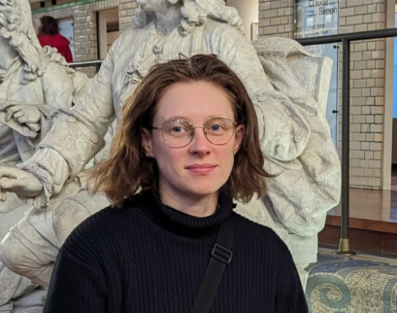 A photo of Eli, a white nonbinary person in their late 20s. They have chin length brown hair and wear round glasses. They wear a chunky black roll neck jumper and a black crossbody bag. They stand in front of a row of marble statues, on display in La Piscine Museum Roubaix.