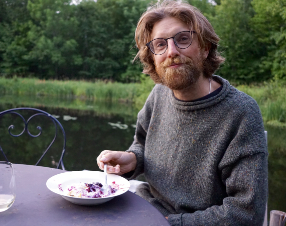 A shaggy looking thirty year old eating pie in front of a lake