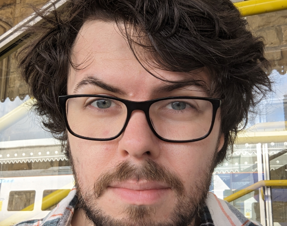 Picture of Adam Clewes-Boyne, a white man with dark brown hair, black glasses, a moustache, and beard