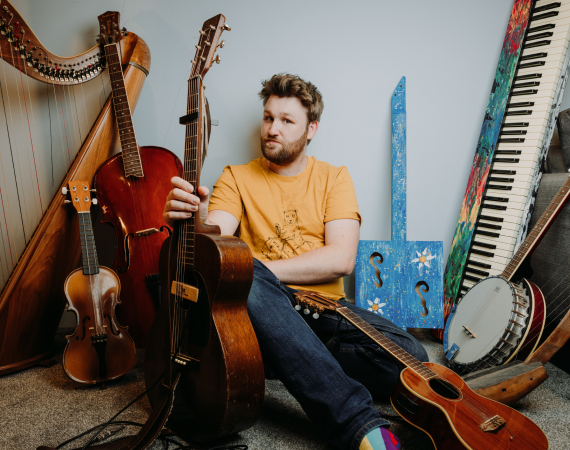 Greg wearing a yellow t-shirt sitting in amongst several musical instruments including a harp, piano, guitar and banjo