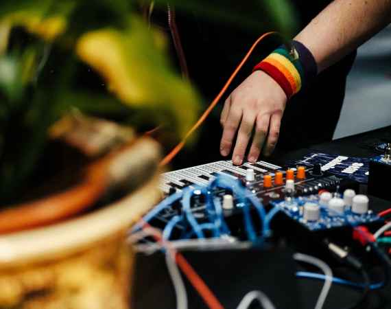 A photo of a hand wearing a rainbow wristband touching a key on a synthesizer surrounded by audio cables and guitar pedals. in the foreground there is an out of focus plant in a giant pot.