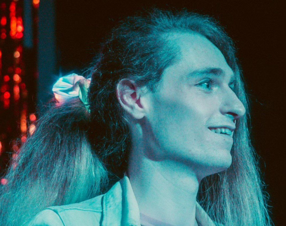 Heidi, a white nonbinary person with long brown hair tied up in an LED scrunchie, is smiling on stage in blue lights.